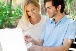 Young couple working on laptop and smiling while sitting relaxed on bench at summer park